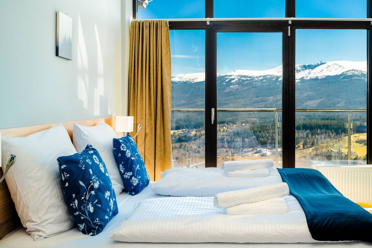Un dormitorio con un amplio ventanal, una cama con ropa de cama blanca y vistas a la montaña.