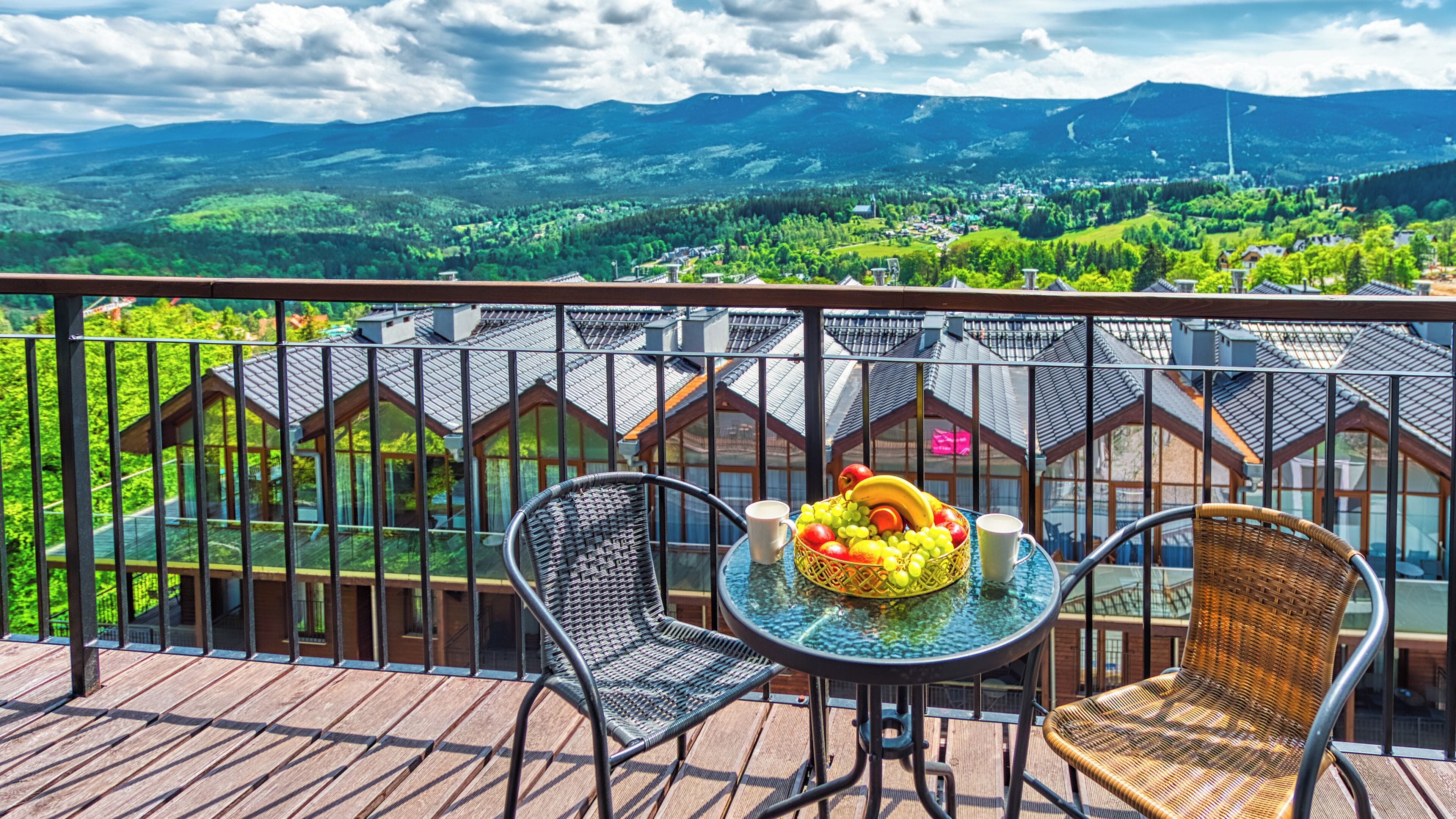 A balcony with wicker chairs, a table with fruit and a cup, and a view of a village and mountains.