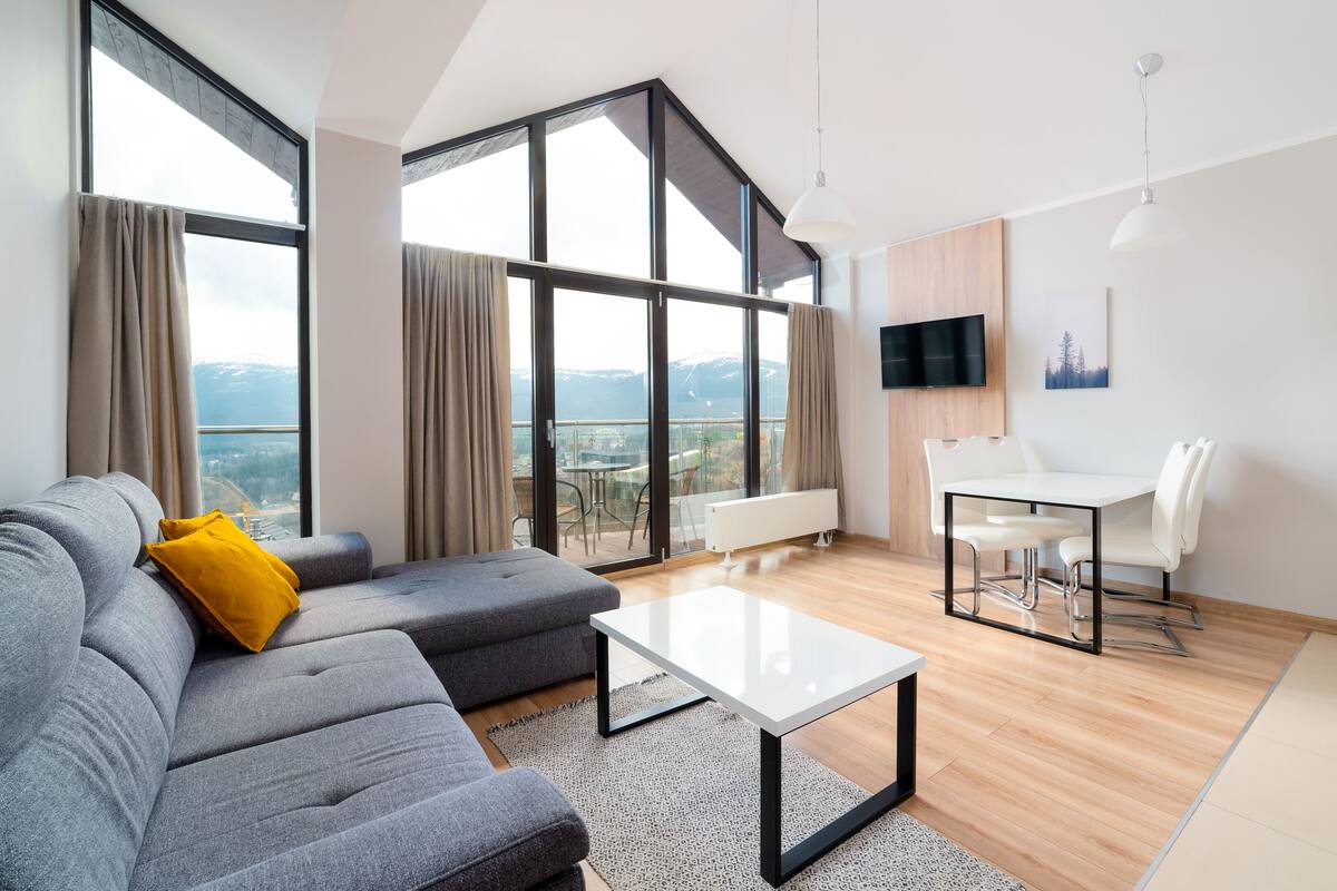 Una sala moderna con un sofá gris, una mesa de centro blanca y un comedor con una mesa y sillas blancas. Amplias ventanas ofrecen vistas a un paisaje montañoso.