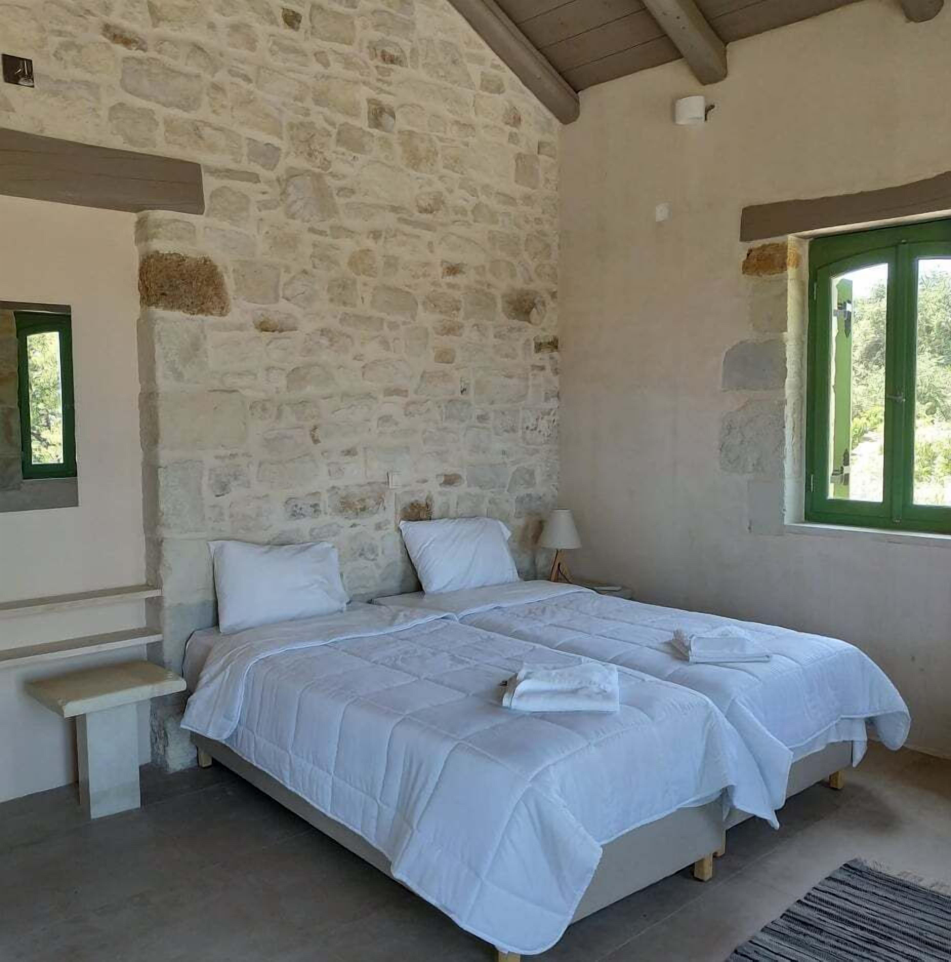 Quarto com parede de pedra, cama com roupa de cama branca, mesa de cabeceira e janela com moldura verde.