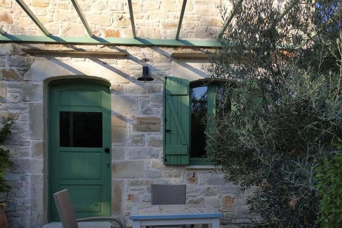 Un edificio de piedra con puerta verde y contraventanas, una mesa con sillas y plantas en macetas.