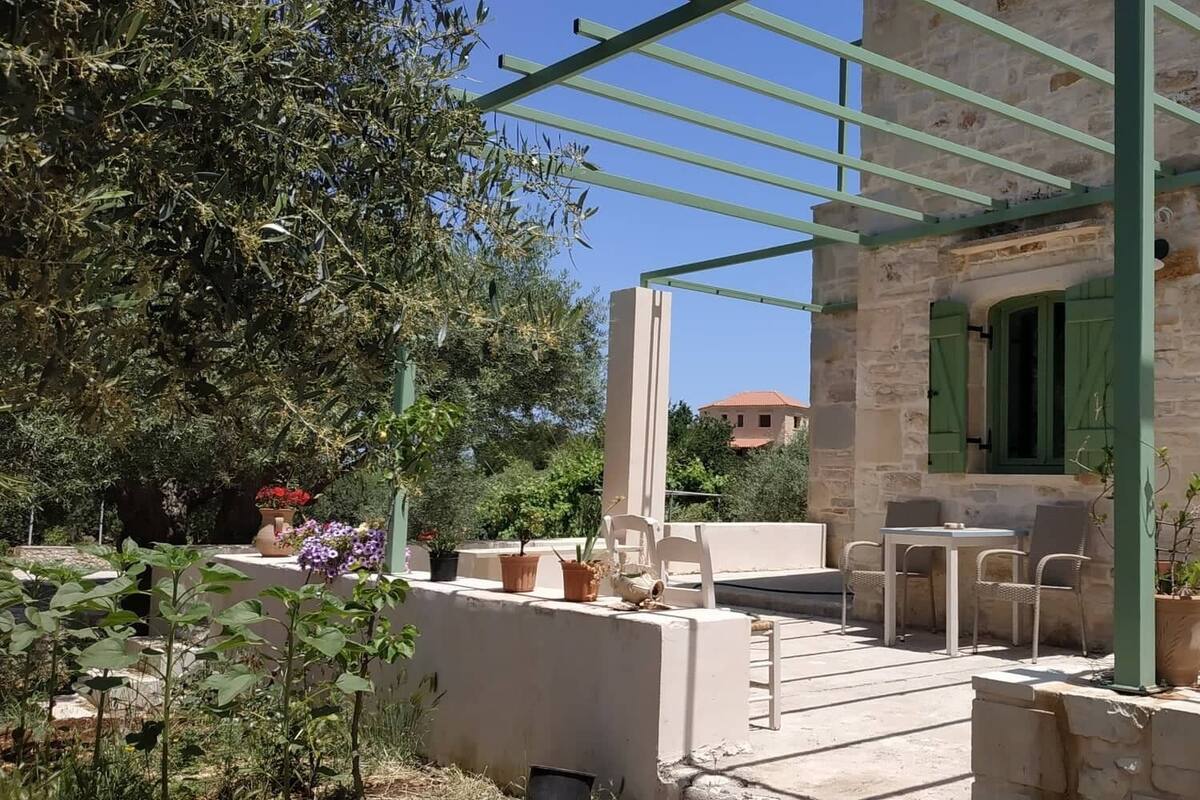 Un edificio de piedra con puerta y ventanas verdes, un porche cubierto con mobiliario exterior y plantas en macetas.