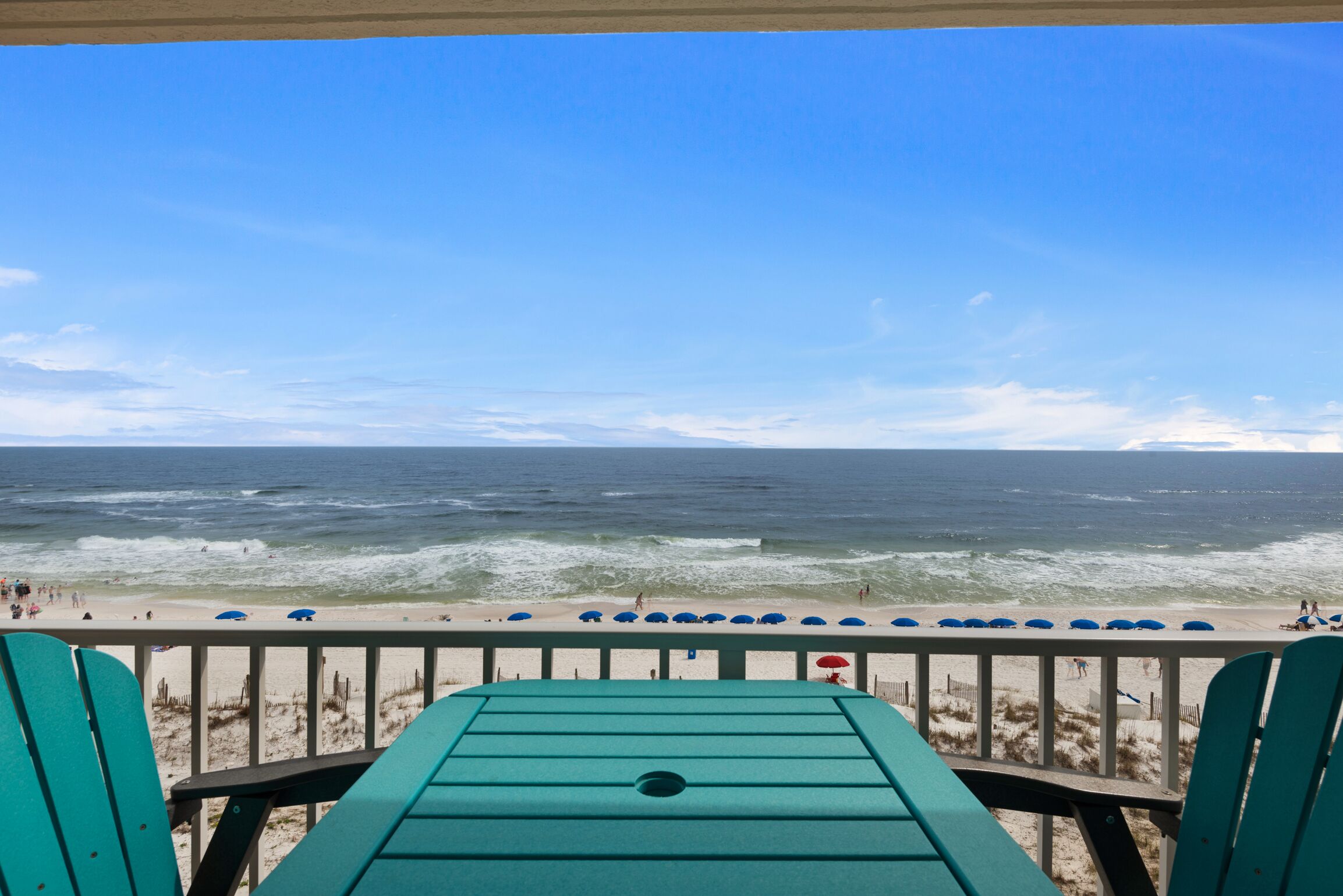 A beach view from a covered deck with a railing, overlooking the ocean and beachgoers.