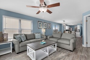 A living room with two grey sofas, a coffee table, and a ceiling fan. There are three framed pictures on the wall and a window with blinds.