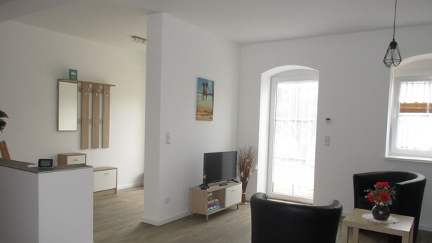 A modern living room with a flat-screen TV, a black armchair, a small wooden table with a vase, and a large window providing natural light. - Vacation apartment "Zum alten Pferdestall" - Somewhere in the middle of nowhere (Zichow)