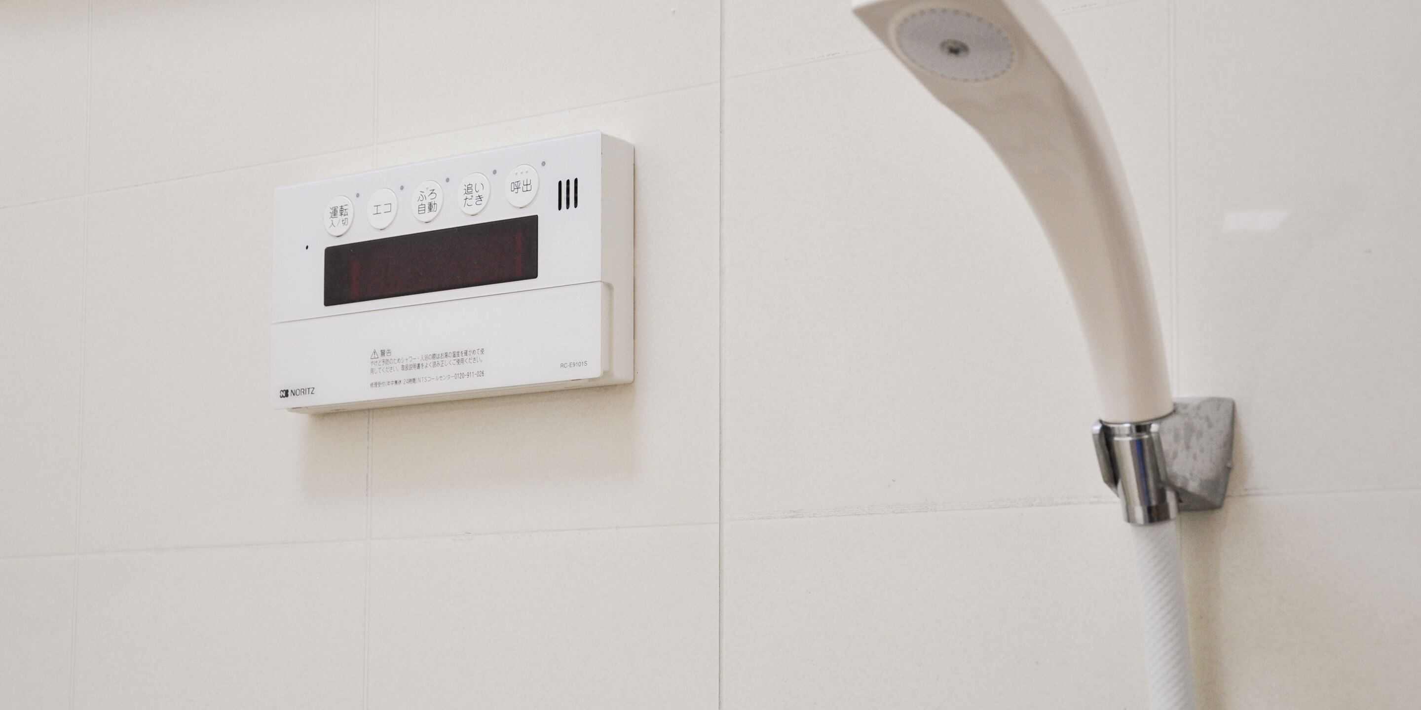 Panel de control de ducha blanco con texto en japonés y pantalla digital, fijado a una pared con azulejos.