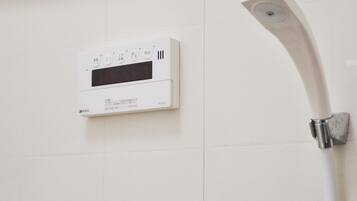 Panel de control de ducha blanco con texto en japonés y pantalla digital, fijado a una pared con azulejos.