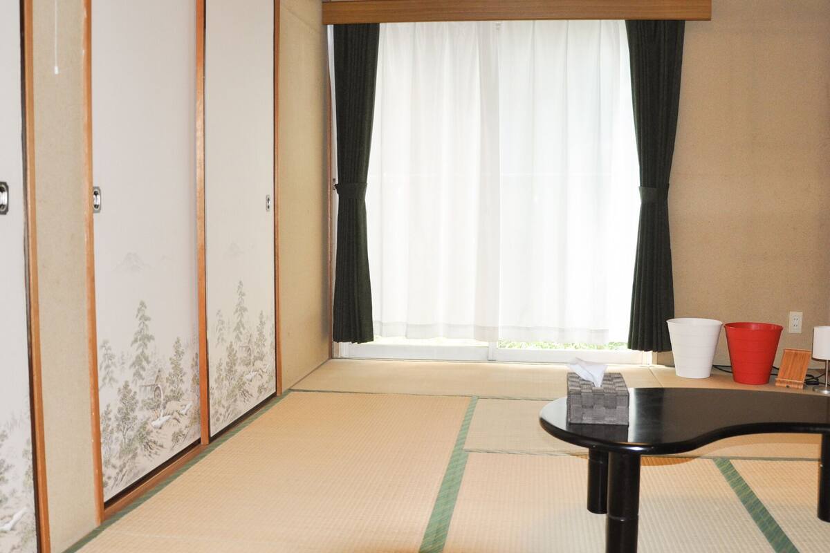 A traditional Japanese room with tatami flooring, a sliding shoji door, a futon bed, and a small table with cups and a tissue box.