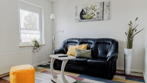 Una sala de estar moderna con un sofá de cuero negro, una mesa de centro blanca, un tapete a rayas de colores, un otomano naranja pequeño y una planta en maceta.