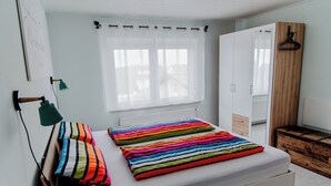 Un dormitorio con cama, mesita de noche, lámpara, ventana con persianas, armario y cómoda de madera.