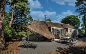 Et stråtækt hus med en patio, herunder et bord og stole, omgivet af træer.
