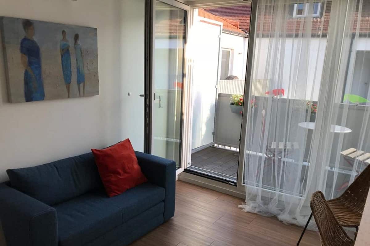 Ein Zimmer mit einem blauen Sofa, einem roten Kissen, einer Glastür zu einem Außenbereich mit Tisch und Stühlen sowie einem Bild an der Wand.