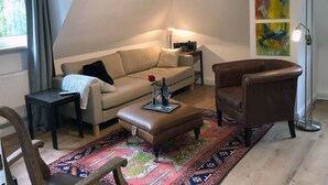 Een gezellige woonkamer met een bank, fauteuil, voetenbankje, een kleine tafel, een vloerlamp en een kleurrijk vloerkleed.