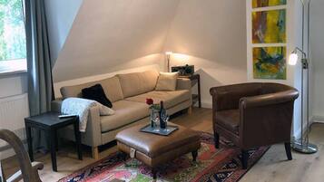 Ein gemütliches Wohnzimmer mit Sofa, Sessel und Hocker, einem kleinen Tisch, einer Stehlampe und einem farbenfrohen Teppich.