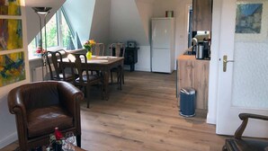 Een eetgedeelte met een houten tafel en stoelen, een kleine prullenbak, en een koelkast op de achtergrond.