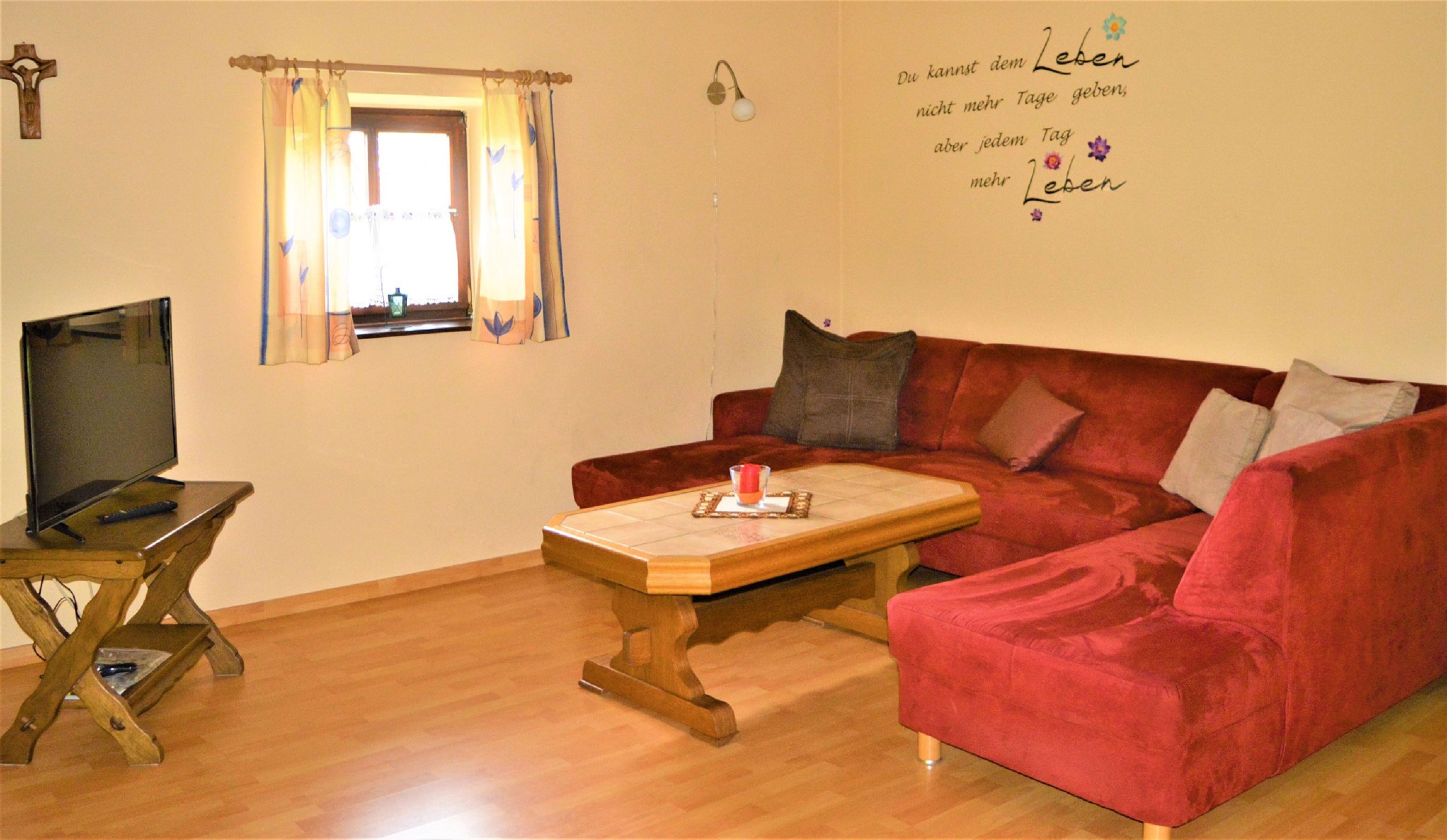 Ein gemütliches Wohnzimmer mit einem roten Sofa, einem Holztisch, einem Fernseher auf einem Holzständer und einem Fenster mit Vorhängen.