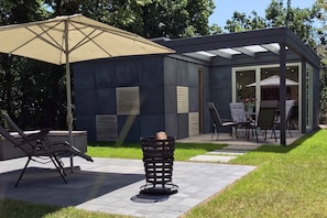 A modern outdoor patio with a covered seating area, a fire pit, and a glass-enclosed dining space. - Cube-House Ferienhaus in Franken (Volkach)