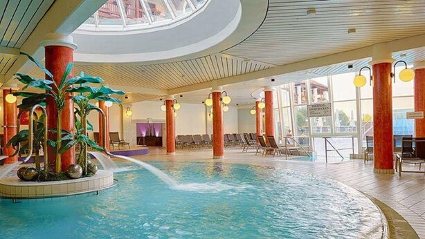 An indoor swimming pool with a water feature, surrounded by lounge chairs and a large window.