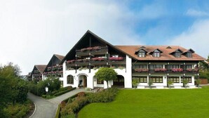 A large, multi-story hotel with a traditional Alpine architectural style, featuring balconies with flower boxes and a well-maintained lawn.