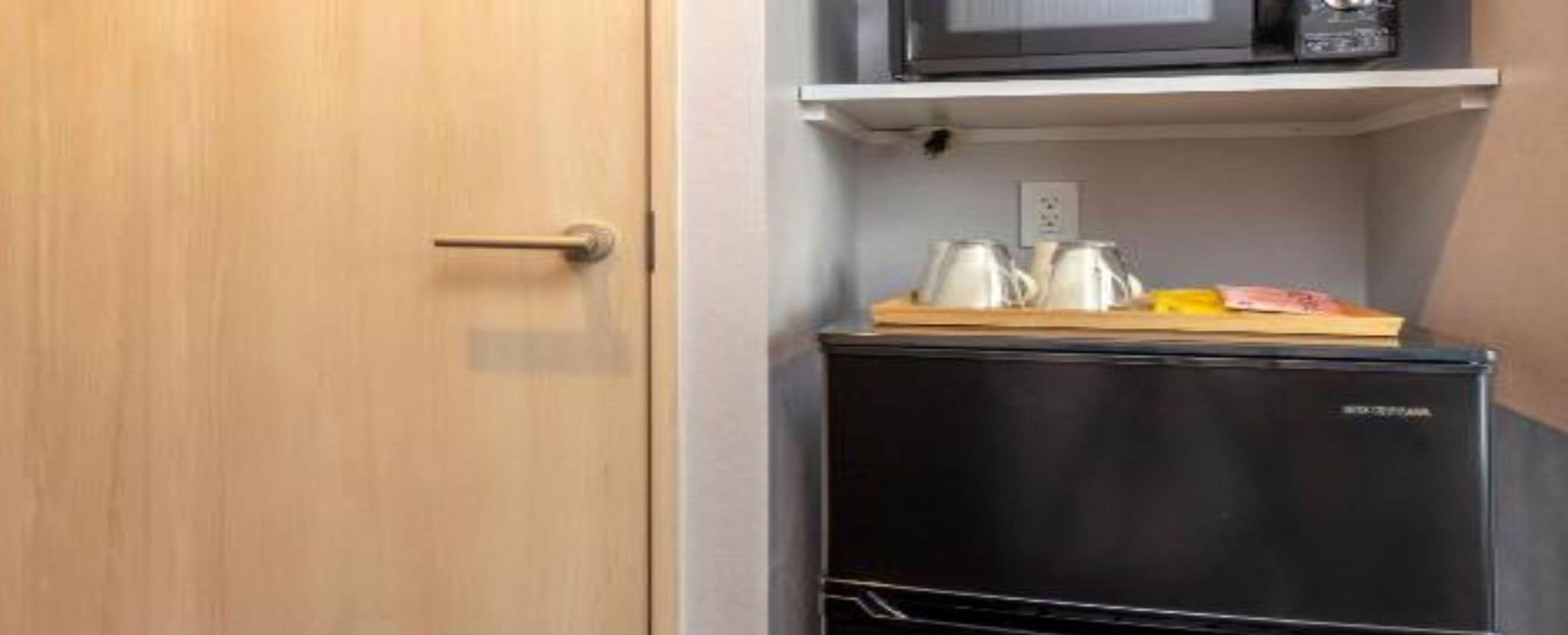 A compact kitchenette with a microwave, a small refrigerator, and a shelf with kitchenware.