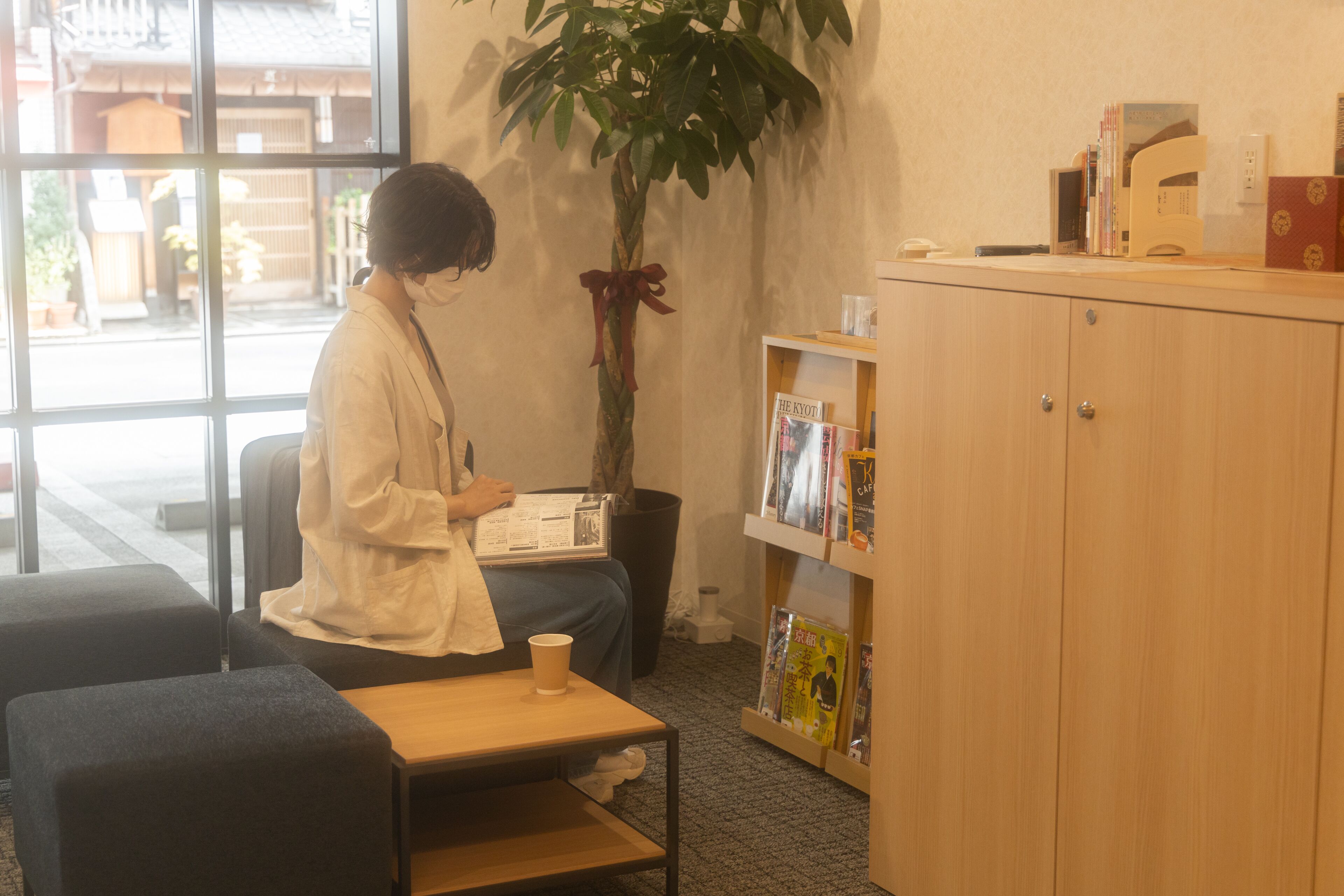 A person seated at a table with a cup, a bookshelf with books, and a potted plant in a room with a window and a wooden cabinet.