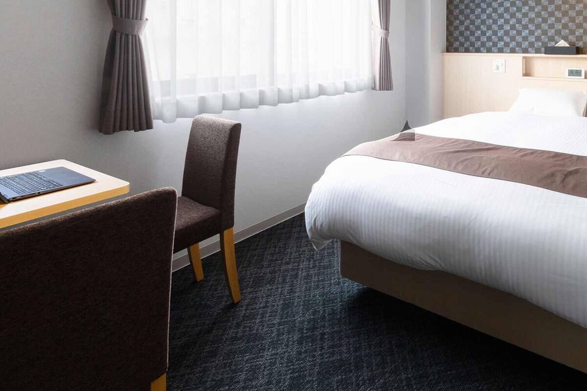 Une chambre d’hôtel avec un lit, un bureau avec un ordinateur portable, une chaise et une fenêtre avec des rideaux.