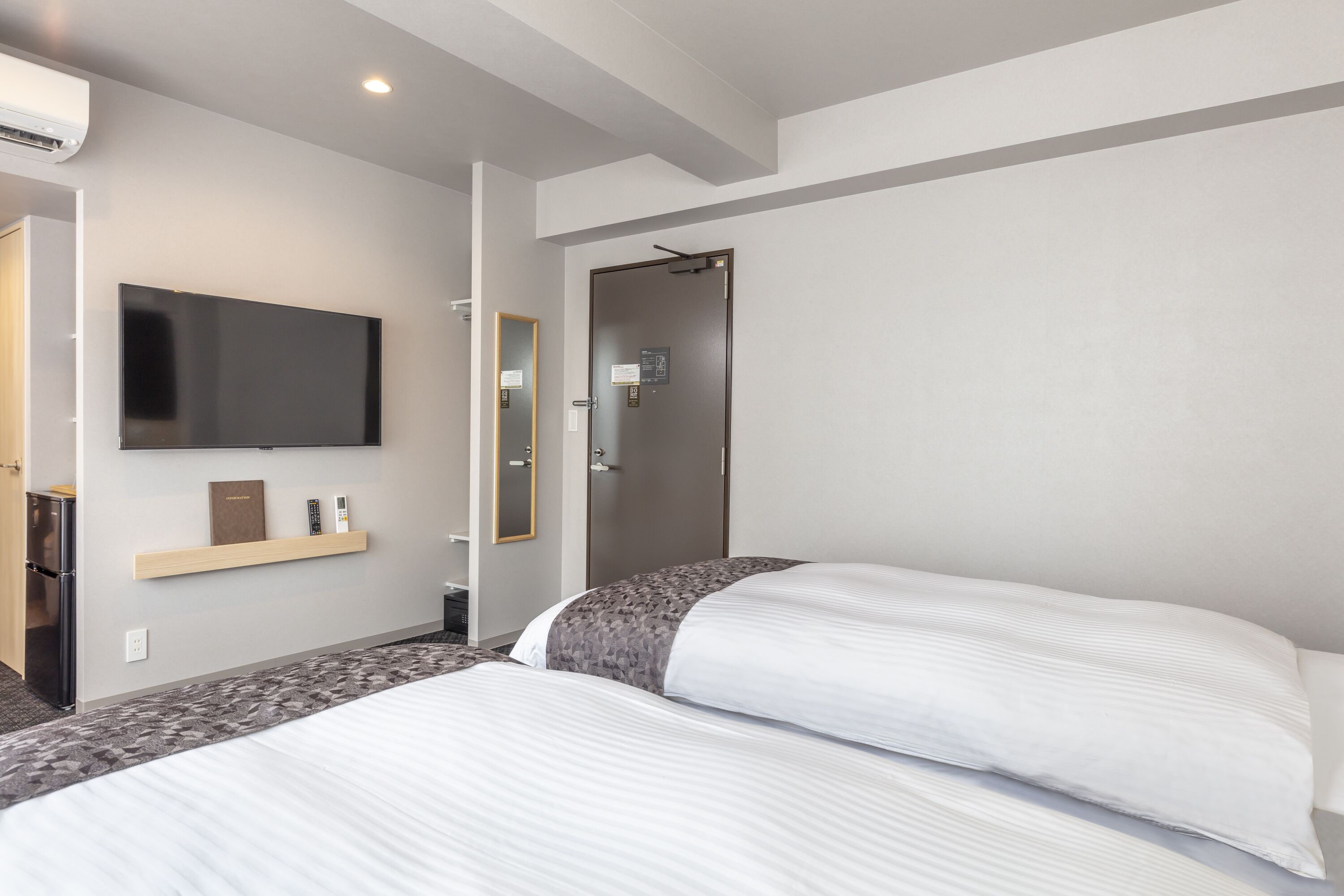 A modern hotel room with a large bed, a flat-screen TV mounted on the wall, a small desk with a lamp, and a door leading to a bathroom.