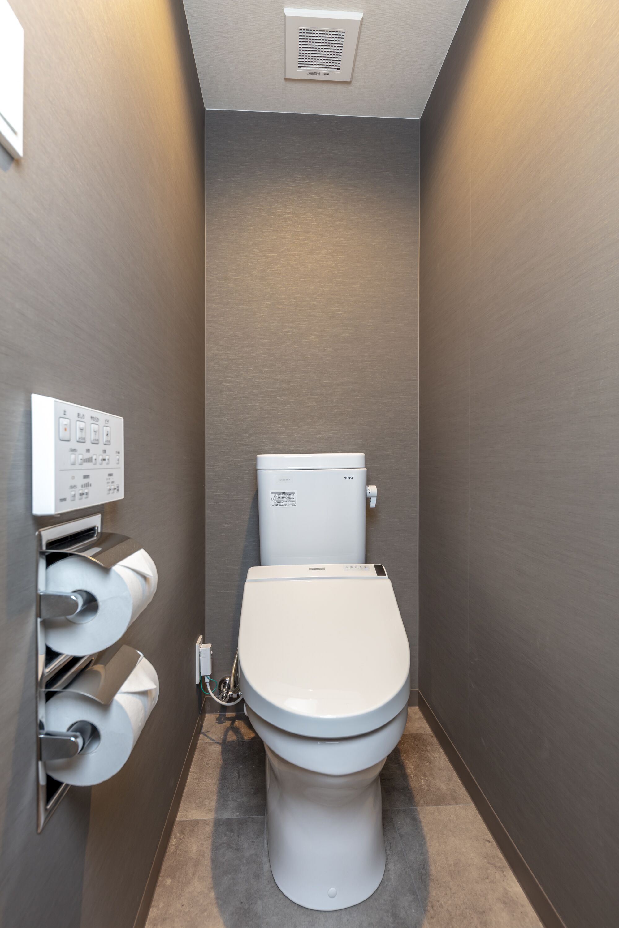 A compact bathroom with a toilet, a wall-mounted toilet paper dispenser, and a vent on the ceiling.