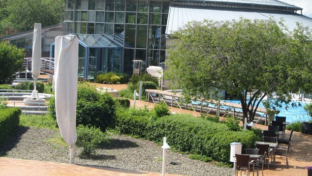 A modern building with a glass facade and a metal roof, surrounded by a landscaped garden with a pool and outdoor seating. - Ferienwohnungen an der Therme (Bad Rodach)