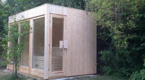 A wooden sauna with a sliding door and a small window, surrounded by dense greenery. - Ferienhaus Sine Tempo (Marktleugast)