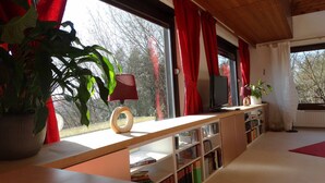 A room with a large window, red curtains, a wooden desk, a television, and bookshelves. - Ferienhaus Sine Tempo (Marktleugast)