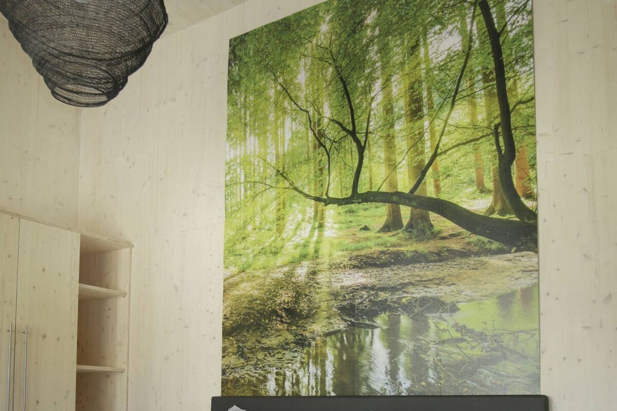 A bedroom with a large bed, a headboard, a wardrobe, and a wall-mounted painting of a forest scene.