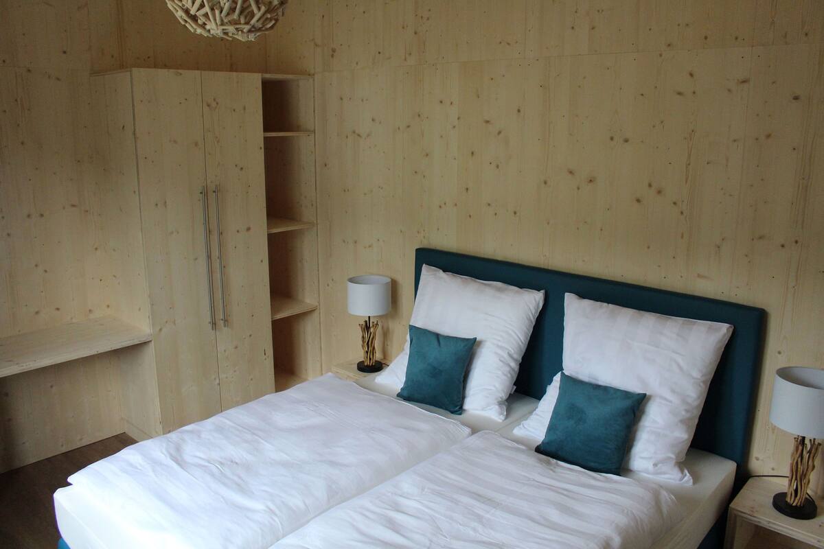A double bed with white linens and teal pillows, a wooden wardrobe, and a bedside table with a lamp.