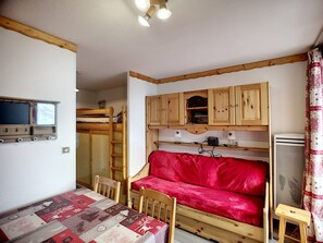 A compact room with a bunk bed, a red sofa, a wooden table, and a small kitchenette.