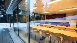 A conference room with multiple tables and chairs, large windows, and a view of trees outside.