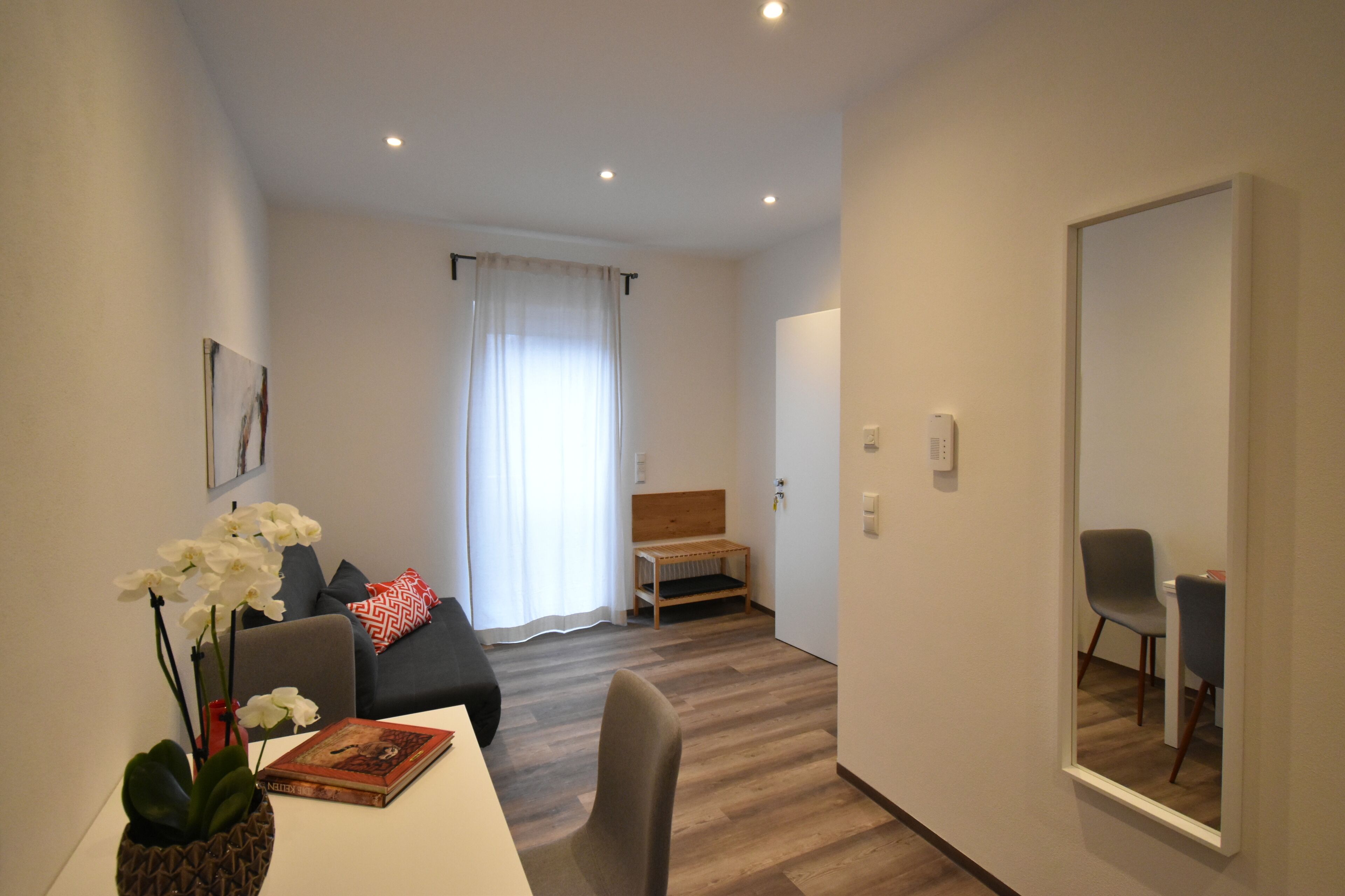 A modern hotel room with a white bed, a small wooden bench, a grey armchair, and a dining table with a vase of white flowers.