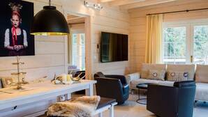 A modern living room with a white wooden wall, a black leather sofa, a white dining table with a fur throw, and a mounted TV. - Chalet Bischofsmühle (Helmbrechts)