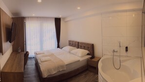 Una habitación de hotel moderna con una cama grande, un escritorio de madera, un televisor de pantalla plana, ducha y un gran ventanal con cortinas.