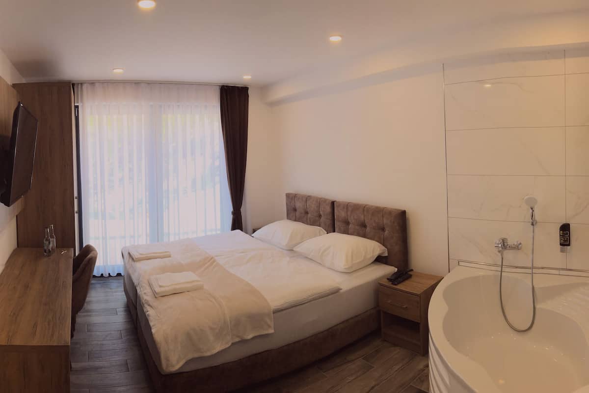Una habitación de hotel moderna con una cama grande, un escritorio de madera, un televisor de pantalla plana, ducha y un gran ventanal con cortinas.