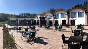 A modern outdoor seating area with tables and chairs, a building with large windows, and a clear sky. - Hotel Asador (Netphen)
