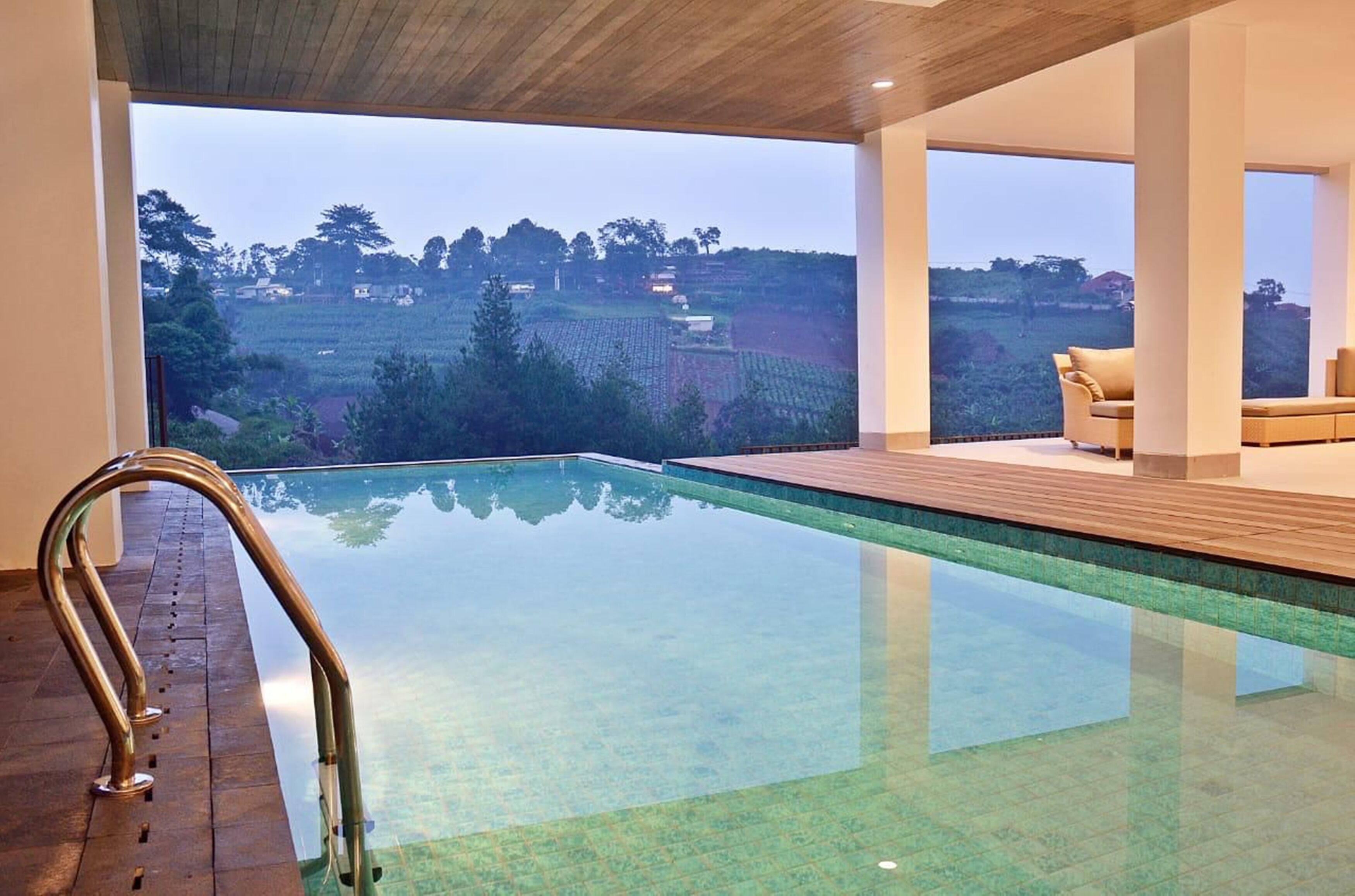 A modern indoor pool with clear glass walls, wooden deck, and a view of a hilly landscape.