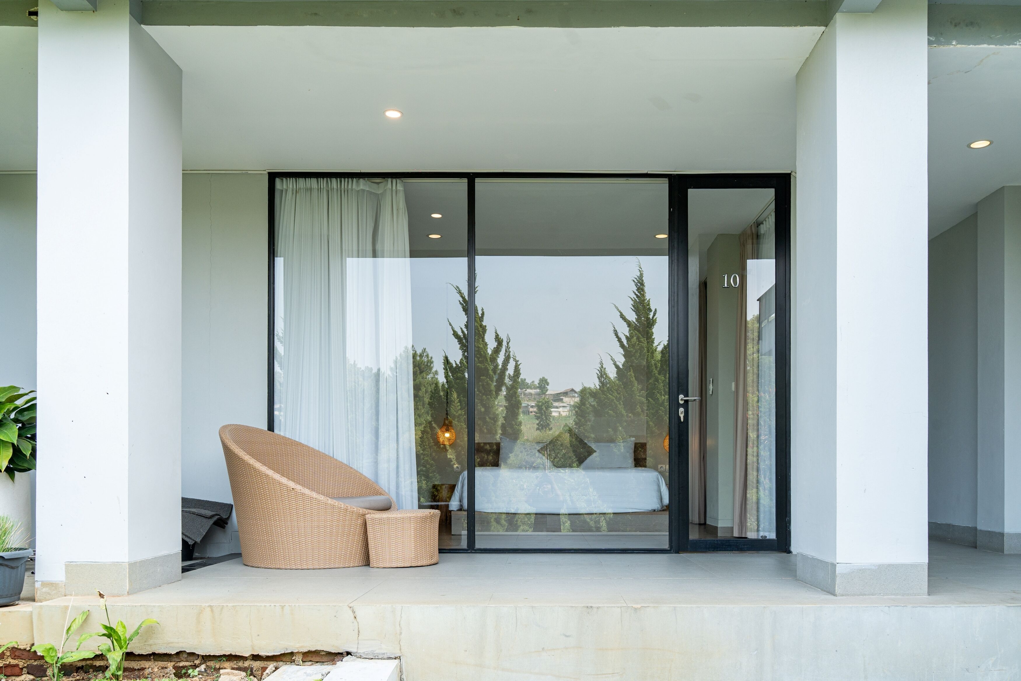 A modern room with a glass door leading to a balcony, a wicker chair, a bed, and a view of trees.