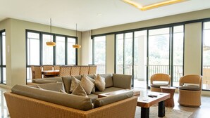 A spacious living area with a large sofa, wicker chairs, a wooden coffee table, and large windows offering a view of the outdoors.