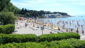 A beach with sun loungers and umbrellas, a sign indicating no swimming, and a view of the sea and buildings in the background. - Apartments Bulovic (Split)