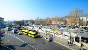 A bus station with multiple buses, including a yellow one, and a few cars. There are people walking and standing around. - Apartments Bulovic (Split)