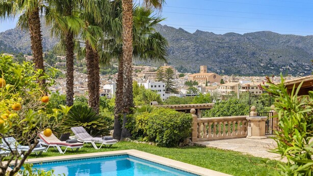 Un área de piscina con sillones, palmeras y vistas a un pueblo con edificios y montañas al fondo.