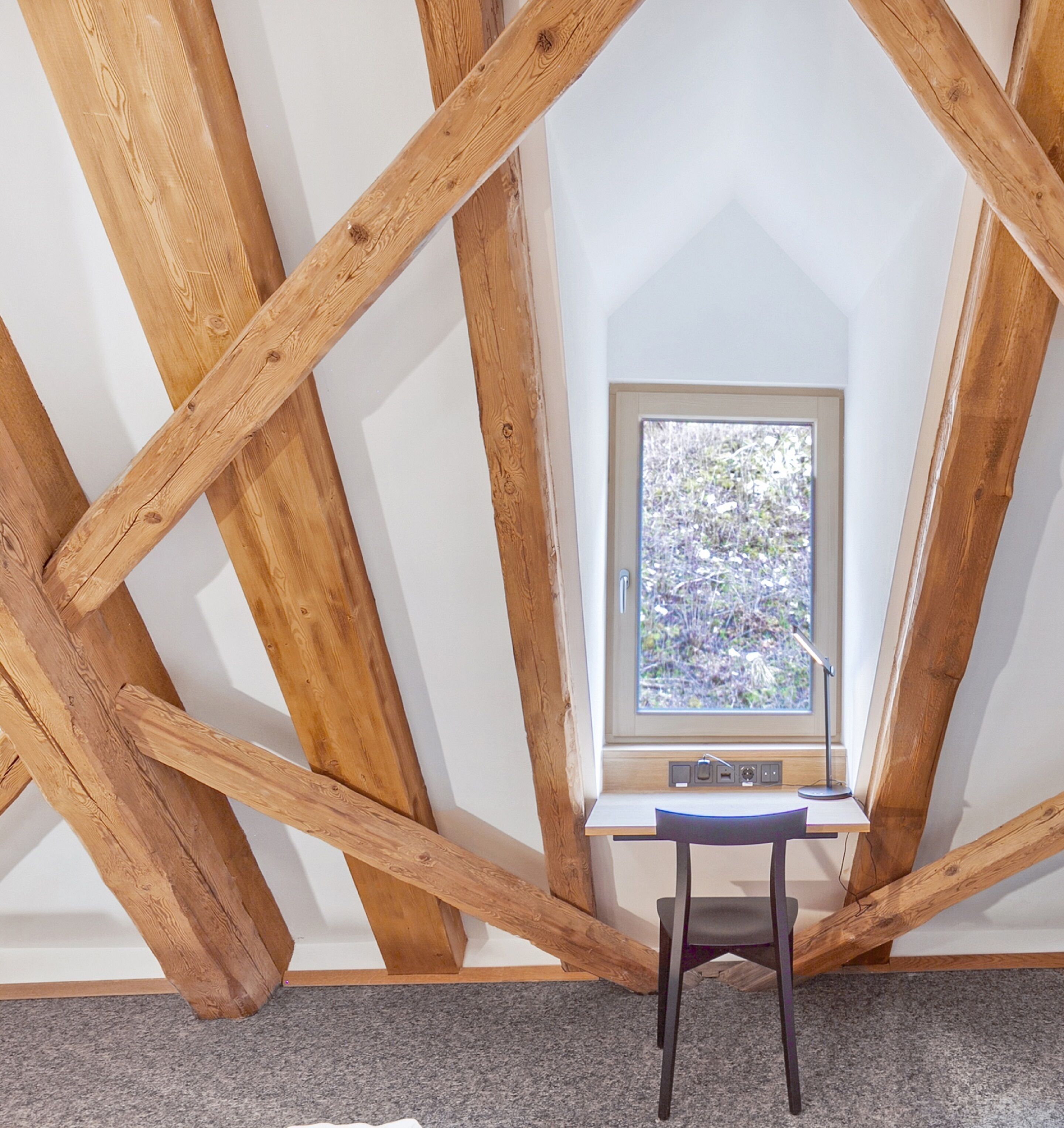 Una habitación con techo inclinado, vigas de madera, un ventanal pequeño y un escritorio con silla.