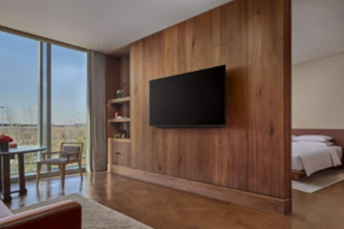Ein modernes Hotelzimmer mit großem Fenster, Holzvertäfelung, einem flachbildfernseher an der Wand, einem Bett mit weißen Bettwäsche und einem kleinen Tisch mit Stühlen.