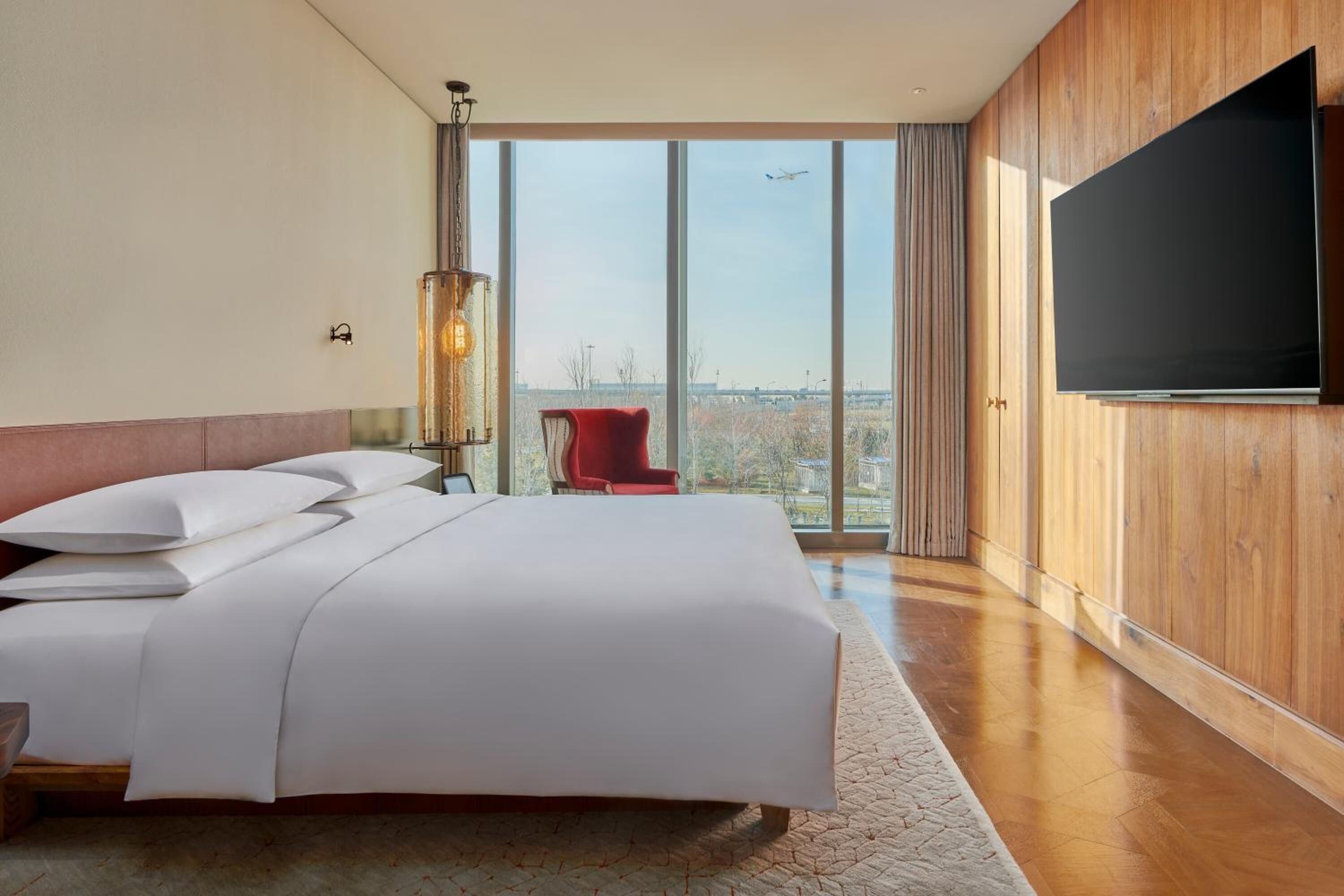 A modern hotel room with a large bed, a TV, a red chair, and a view of the cityscape through large windows.