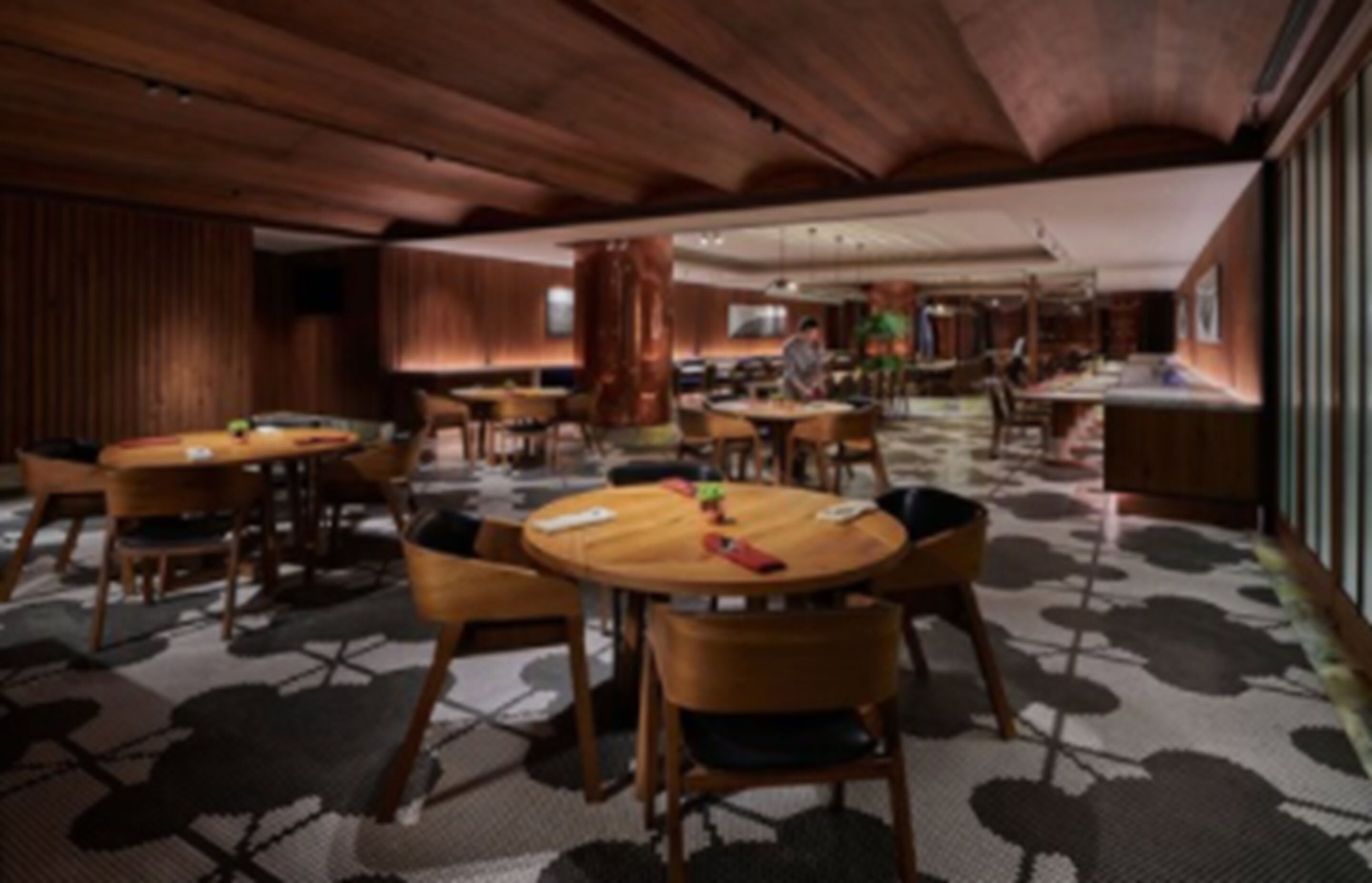 Un vaste espace dédié à la salle à manger, avec des tables et des chaises en bois rondes, une moquette à motifs et un plafond haut orné de poutres en bois.